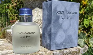 Perfumy Dolce & Gabbana Light Blue Pour Homme, w szklanej butelce i niebieskim pudełku, na kamiennym murku wśród zieleni.