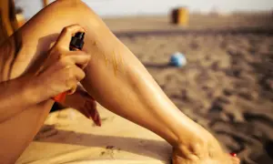 Kobieta aplikuje olejek na nogę, by jak się szybko opalić domowe sposoby. Na plaży w tle widać piasek i piłkę.