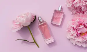 Dwa flakoniki różowych perfum leżą obok delikatnych kwiatów pivoine na pastelowym tle.
