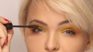Kobieta z blond włosami nakłada tusz do rzęs. Jej oczy podkreślone są żółtym cieniem, idealnym do żółtej sukienki.