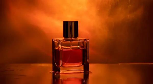 Szklana buteleczka perfum z bursztynowym płynem, otoczona ciepłym, pomarańczowym światłem. Zapach ambrowy unosi się w powietrzu.