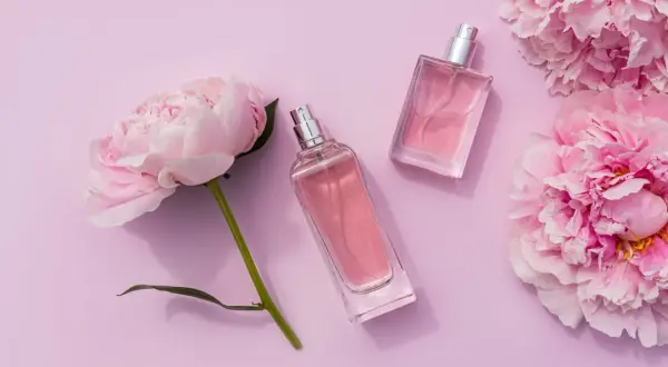 Dwa flakoniki różowych perfum leżą obok delikatnych kwiatów pivoine na pastelowym tle.
