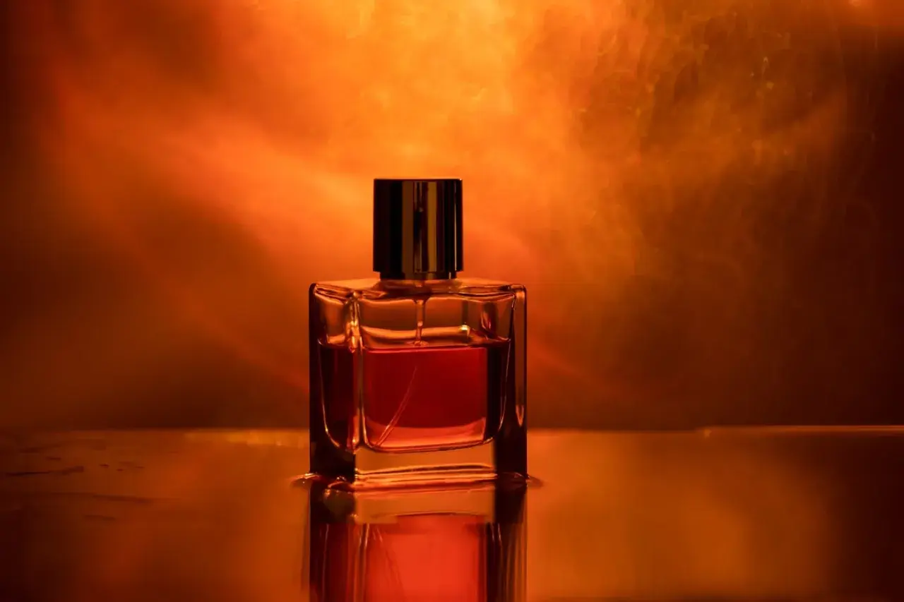 Szklana buteleczka perfum z bursztynowym płynem, otoczona ciepłym, pomarańczowym światłem. Zapach ambrowy unosi się w powietrzu.