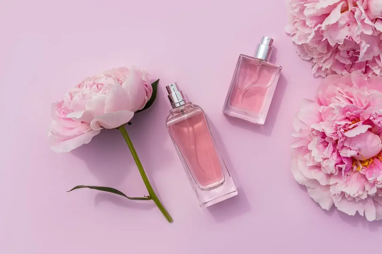 Dwa flakoniki różowych perfum leżą obok delikatnych kwiatów pivoine na pastelowym tle.