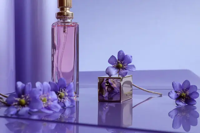 Fiołki otaczają flakon perfum, tworząc subtelny zapach fiołków. Złoty sześcian z kwiatem dodaje elegancji.